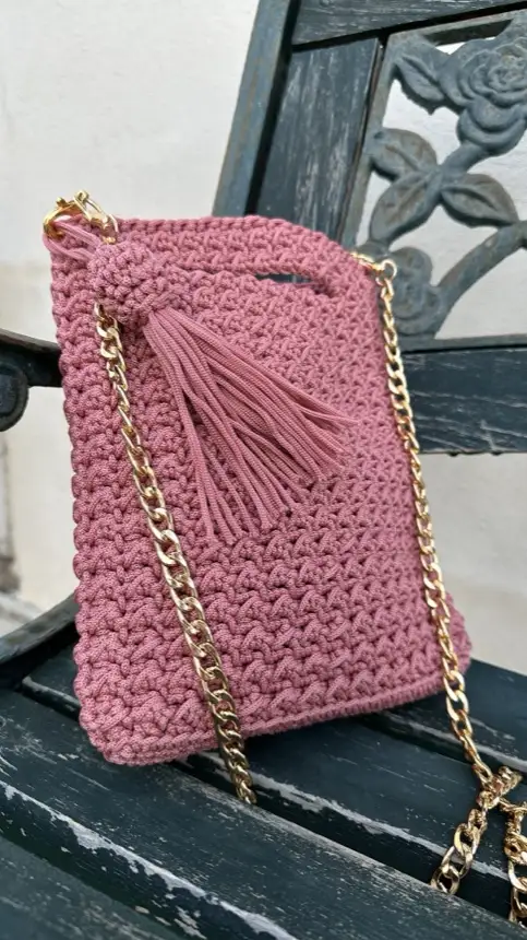 Pochette rosa antico all’uncinetto con catenella dorata e nappa, su una panchina in ferro battuto